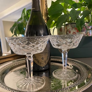 May include: Two clear crystal coupe glasses featuring a diamond-cut pattern on the bowl and a faceted stem. The glasses have a circular base with a sunburst design. They are positioned on a round silver tray in front of a dark glass bottle.