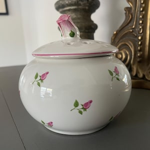 May include: A white ceramic potpourri jar with a rounded body and a removable lid. The jar features a pattern of small pink rosebuds with green leaves. The lid has a sculpted pink rosebud handle and several small circular ventilation holes.