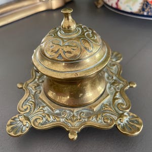 May include: Antique brass inkwell with a domed lid and ornate, floral-inspired detailing. The inkwell sits on a decorative base with a scalloped edge. The brass has a warm, aged patina, suggesting its vintage origin. A classic desk accessory.