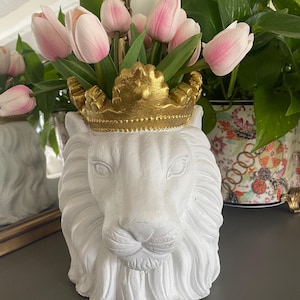 May include: A white lion head planter with a gold crown holding pink tulips. The planter is made of a textured material and has detailed facial features. The tulips have green stems and leaves.