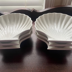 May include: A set of white ceramic shell-shaped dishes. Each dish has a fluted design radiating from the center. The dishes are stacked in pairs, showcasing their elegant, coastal-inspired design. Perfect for serving appetizers or desserts.