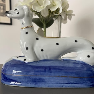 May include: A white ceramic dog figurine with black spots, a gold-painted collar, and a blue-painted base. The dog is in a reclining position, facing left, with a smooth, glossy finish and gold trim detailing along the edge of the blue base.