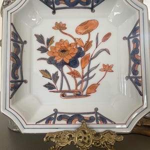May include: A white porcelain dish with a square shape and a decorative floral design. The dish features a central orange and blue floral motif, surrounded by blue and red accents. The dish is supported by a gold-colored metal stand.