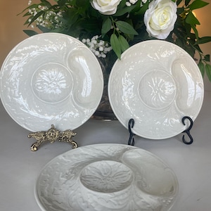 May include: Three white ceramic serving dishes with floral embossed designs. Two dishes are round with two side compartments and a central floral design. The third dish is oval with a central dip bowl and two side compartments. Decorative servingware.