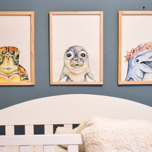 May include: Three framed watercolor paintings of sea creatures. The first painting features a turtle with a green, brown, and yellow shell. The second painting features a seal with a gray and white coat. The third painting features a dolphin with a blue and gray body and a pink and white flower crown.