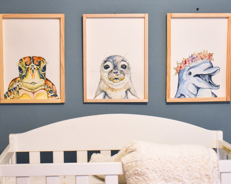 May include: Three framed watercolor paintings of sea creatures. The first painting features a turtle with a green, brown, and yellow shell. The second painting features a seal with a gray and white coat. The third painting features a dolphin with a blue and gray body and a pink and white flower crown.