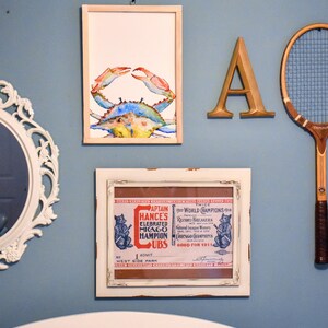 May include: A framed watercolor painting of a blue crab on a light blue wall. The painting is surrounded by other framed artwork, including a vintage baseball ticket and a letter A.