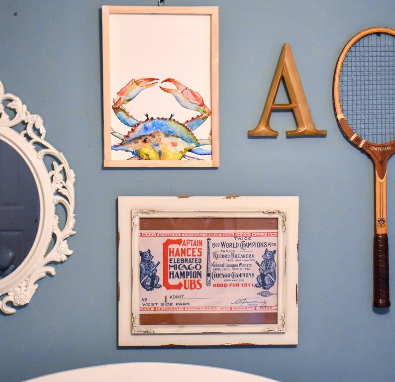 May include: A framed watercolor painting of a blue crab on a light blue wall. The painting is surrounded by other framed artwork, including a vintage baseball ticket and a letter A.