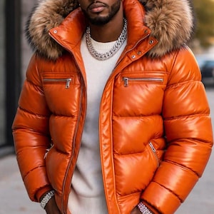 Puede incluir: Una chaqueta acolchada naranja con capucha ribeteada de piel. La chaqueta tiene un aspecto brillante, similar al cuero, con bolsillos con cremallera. La persona lleva gafas de sol y un collar de cadena plateada.