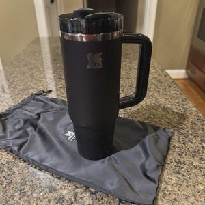 May include: A matte black Stanley tumbler with a matching black handle and a black lid featuring a rotating closure. The Stanley logo is embossed on the upper front of the tumbler. A black drawstring storage bag with the Stanley logo is placed underneath.