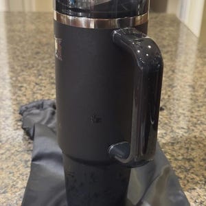 May include: A matte black insulated tumbler with a large side handle, a clear plastic lid with a sliding closure, and a metallic silver rim. The body of the cup features subtle embossed paw print designs and a small logo near the top rim.