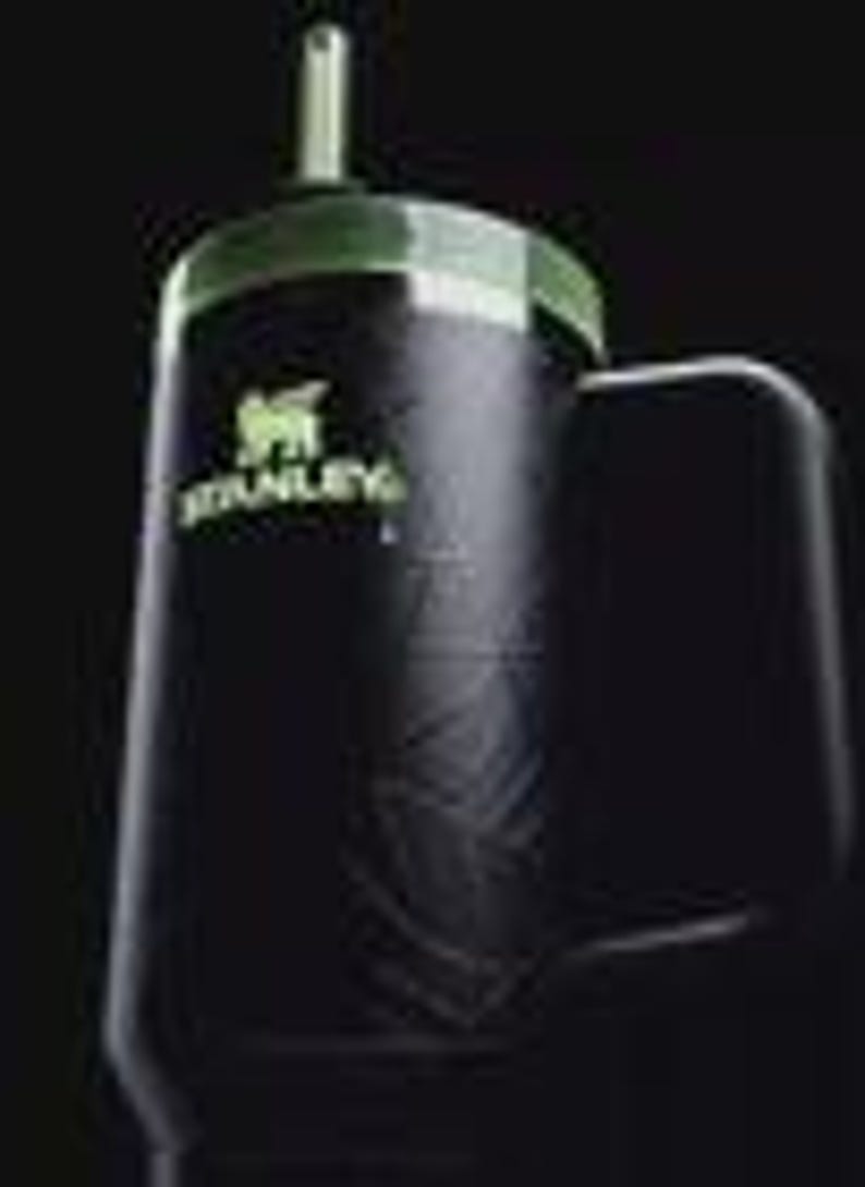 Puede incluir: Un vaso Stanley negro con acabado mate, que presenta una tapa verde y un logotipo verde a juego en la parte frontal. La taza incluye un asa lateral resistente y una pajita que sobresale por la parte superior, con un sutil patr&oacute;n de hojas.