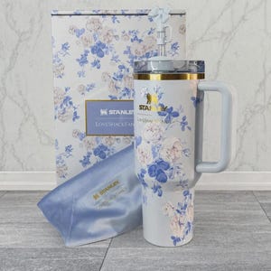 May include: A light blue Stanley tumbler with a white and blue floral pattern, a gold rim, a matching handle, and a straw with a bow topper. It is accompanied by a matching floral box and a light blue satin pouch, both featuring Stanley LoveShackFancy branding.