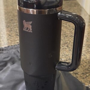May include: A matte black insulated tumbler with a large side handle and a clear plastic lid. The Stanley logo, featuring a winged bear, is printed in a metallic finish on the upper front. The cylindrical body tapers toward the base.