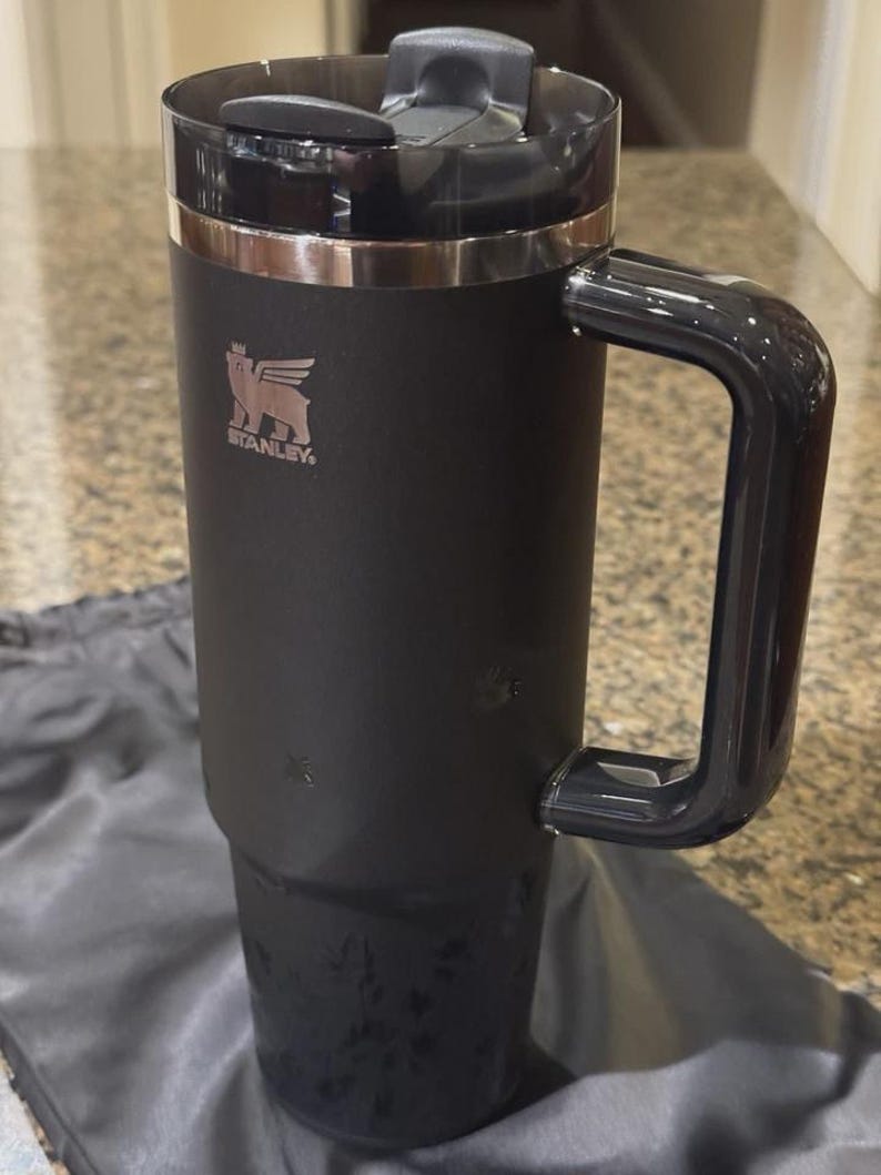 May include: A matte black insulated tumbler with a large side handle and a clear plastic lid. The Stanley logo, featuring a winged bear, is printed in a metallic finish on the upper front. The cylindrical body tapers toward the base.