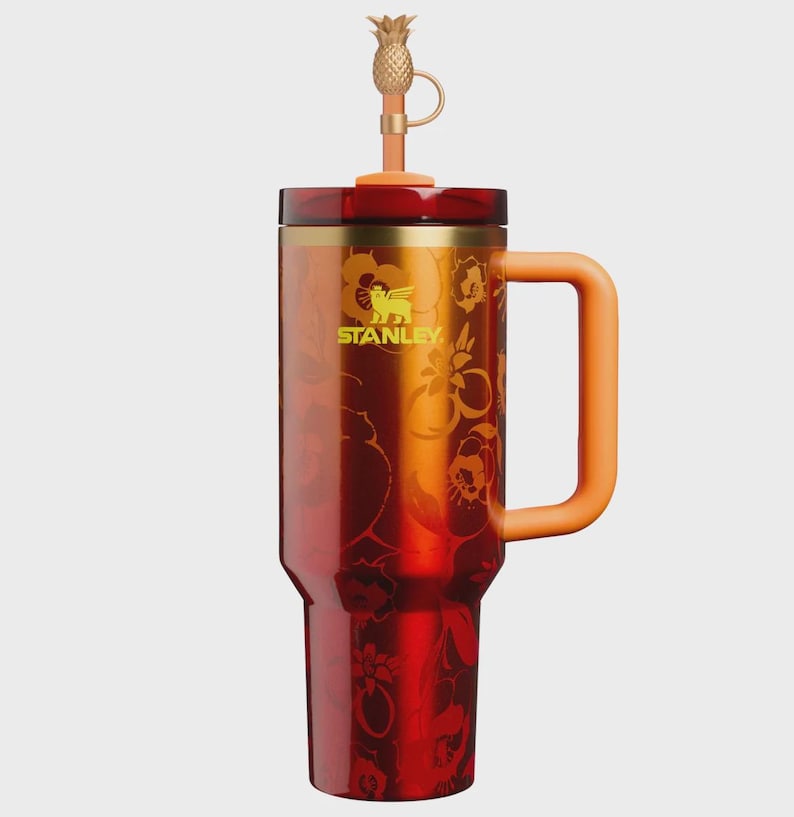 May include: A tall Stanley tumbler with a metallic red finish featuring a tonal floral pattern. It has a gold-toned rim, a solid orange handle, and a matching orange lid. A gold-colored pineapple-shaped topper is attached to the straw.