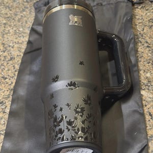 May include: A matte black Stanley tumbler with a large side handle and a black lid featuring a straw. The body of the cup is decorated with a glossy black floral pattern that starts at the base and fades upward. The Stanley logo is printed in silver.