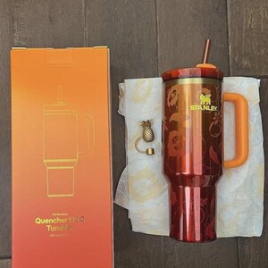May include: A 40 oz Stanley Quencher H2.0 FlowState tumbler in a deep red and orange floral pattern with a matching orange handle, lid, and straw. Beside it is a gold-toned pineapple-shaped straw topper and the original orange gradient box.