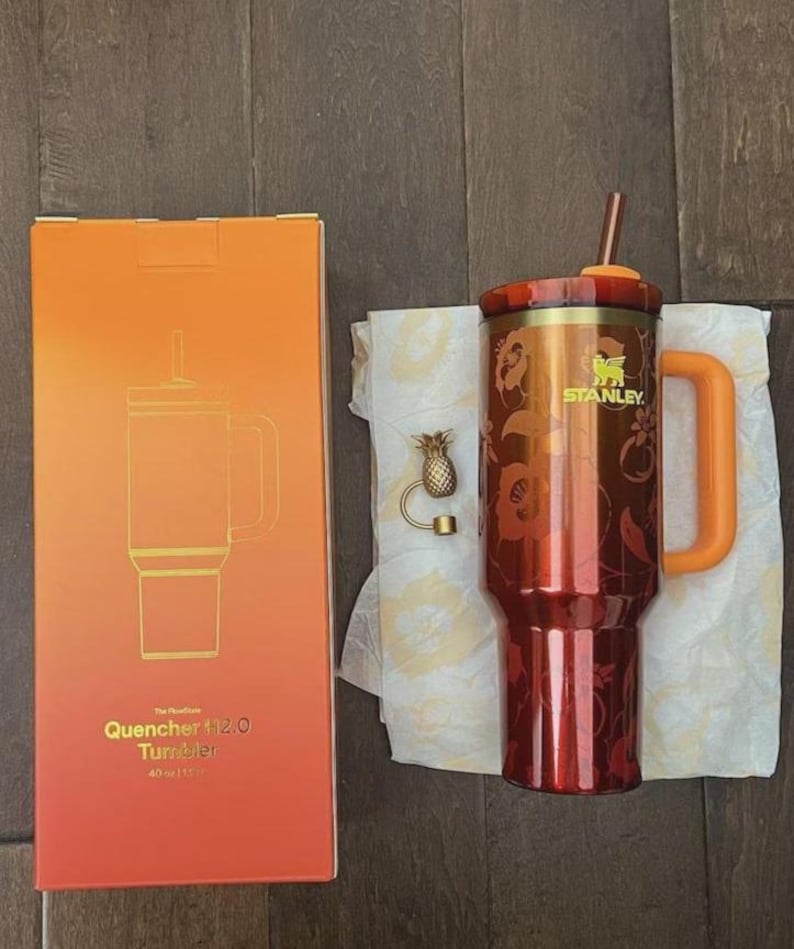 May include: A 40 oz Stanley Quencher H2.0 FlowState tumbler in a deep red and orange floral pattern with a matching orange handle, lid, and straw. Beside it is a gold-toned pineapple-shaped straw topper and the original orange gradient box.