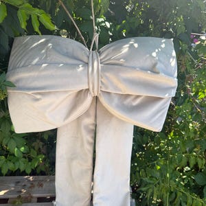 May include: A large, light beige decorative bow made of a smooth, satin-like fabric. The bow features two wide, voluminous loops gathered in the center by a matching fabric band, with two long, straight fabric tails hanging down from the knot.
