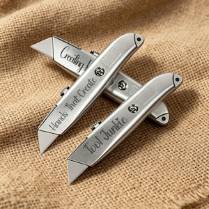 Könnte beinhalten: Zwei silberfarbene, einziehbare Universalmesser mit Metallgehäuse. Ein Messer ist mit dem Text "Hands That Create" graviert, das andere mit "Tool Junkie". Beide Messer verfügen über freiliegende Metallklingen und sichtbare Schrauben.