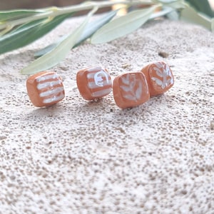 May include: Four small, square-shaped stud earrings with a smooth, terracotta-colored finish. Each earring features a unique, hand-painted white design, including horizontal stripes, a curved line, and delicate leaf patterns.