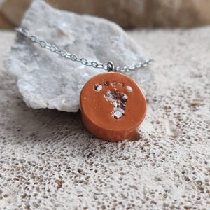 Può includere: Un ciondolo rotondo color terracotta su una catenina in metallo argentato. Il ciondolo presenta un design a impronta incavato riempito con piccole particelle bianche granulari, creando un contrasto materico sulla superficie liscia.