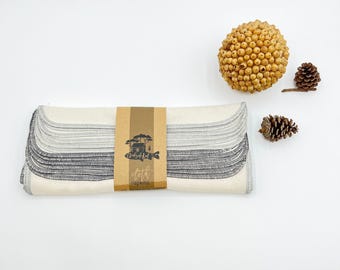 May include: A stack of off-white cloth napkins with dark gray serged edges, secured by a brown paper band featuring a black graphic of a house and trees with the text "Creekside Kid" and "cloth napkins" printed on the front.