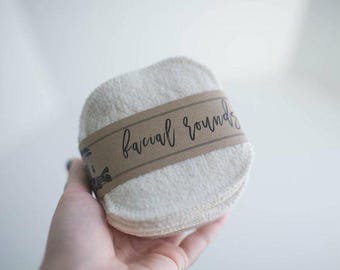 May include: A stack of round, off-white, textured fabric facial pads secured with a brown paper band. The band features the text "facial rounds" printed in a black, cursive script font across the center of the stack.