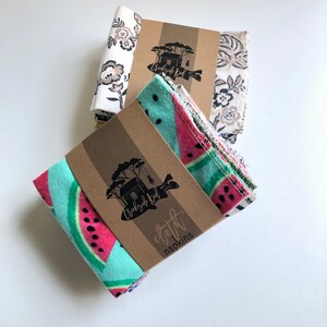 May include: Two stacks of folded cloth napkins secured with brown paper bands. The front stack features a bright turquoise and pink watermelon pattern. The back stack displays a white fabric with a dark brown floral print. Each band features the Creekside Kid logo and text.