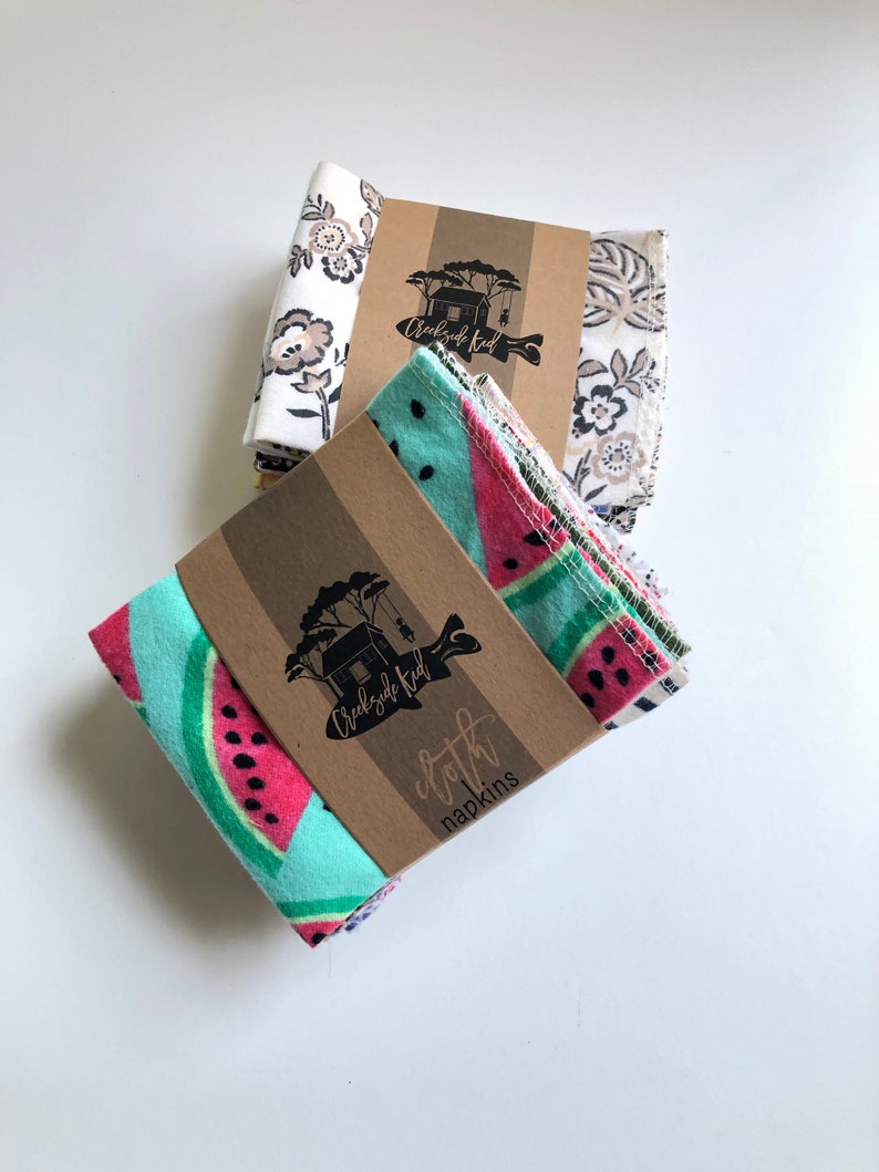 May include: Two stacks of folded cloth napkins secured with brown paper bands. The front stack features a bright turquoise and pink watermelon pattern. The back stack displays a white fabric with a dark brown floral print. Each band features the Creekside Kid logo and text.