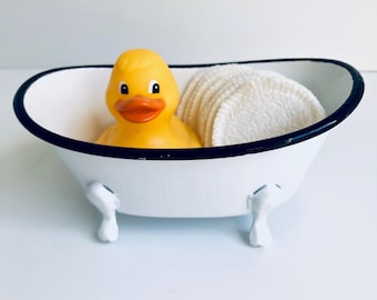May include: A white miniature clawfoot bathtub with a black rim containing a yellow rubber duck and a stack of round white textured cotton facial pads. The small tub features four white feet and a smooth, glossy finish throughout.