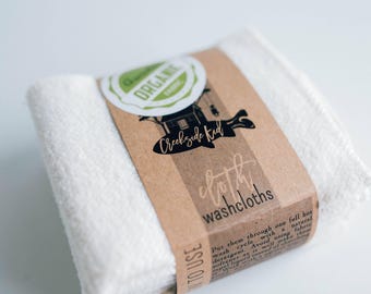 May include: A stack of white textured washcloths wrapped in a brown paper band. The band features a green organic fabric label, a black graphic of a house, and the text Creekside Kid, cloth washcloths, and instructions to use.