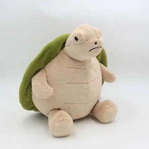 Puede incluir: Un peluche de tortuga con caparazón verde y cuerpo beige. La tortuga tiene una expresión ligeramente gruñona. El juguete está hecho de material suave y está sentado.