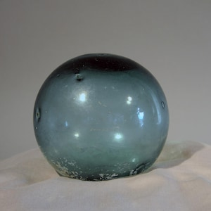 May include: A round, glass float with a dark maroon top and a blue-green body. The glass has a slightly textured surface and small imperfections. The float is resting on a white, textured surface.