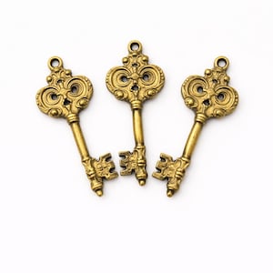 May include: Three antique-style keys with ornate designs. The keys are made of a gold-colored metal and are arranged on a white background. The keys have decorative tops and intricate key bit designs.