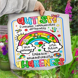 May include: A white tote bag with a colorful design promoting autism awareness. The design includes a rainbow, puzzle pieces, and the words "AUTISM," "Everyone communicates differently," "I love someone with Autism," and "Embrace neurodiversity awareness."
