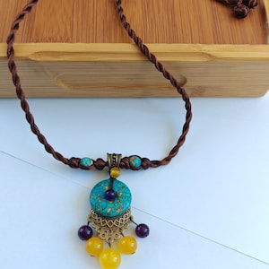 Könnte beinhalten: Eine braune, gedrehte Kordelkette mit einem türkisfarbenen, runden Anhänger mit goldenen Sprenkeln. Der Anhänger ist mit einem bronzefarbenen Filigran-Element, zwei kleinen violetten Perlen und drei gelben Perlen am unteren Ende verziert.