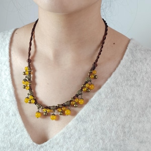 May include: A necklace featuring a dark brown twisted cord adorned with multiple round yellow beads and small metallic bronze-toned accents. The beads are suspended from the cord using decorative bronze-colored caps, creating a layered effect.
