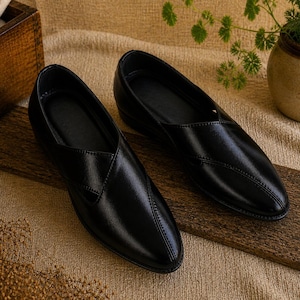 May include: A pair of black leather slip-on shoes with a pointed toe. The shoes are displayed on a wooden plank, with a neutral-toned background. The shoes have a stitched design on the top.