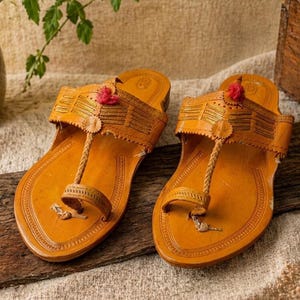 May include: A pair of tan leather sandals featuring a toe post design, intricate stitched patterns on the wide upper strap, and a small red decorative pom-pom accent centered on the top of each sandal, set on a flat, smooth sole.
