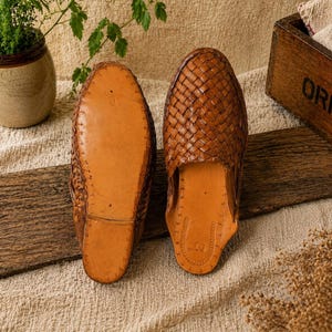 May include: A pair of brown leather woven slip-on shoes. One shoe is shown sole-up, revealing the leather sole. The other shoe is shown right-side up, displaying the woven leather upper. The shoes are on a wooden surface.