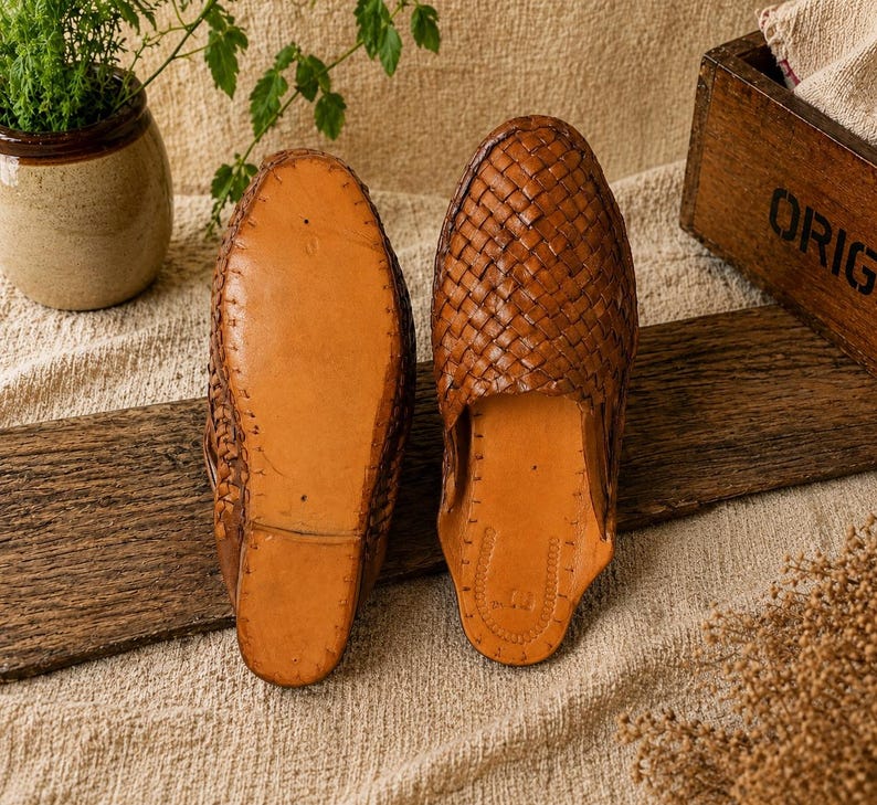 May include: A pair of brown leather woven slip-on shoes. One shoe is shown sole-up, revealing the leather sole. The other shoe is shown right-side up, displaying the woven leather upper. The shoes are on a wooden surface.