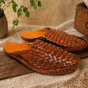 May include: A pair of brown leather woven slippers with a light brown interior. The slippers are displayed on a wooden plank, with a neutral-toned background. The slippers are a slip-on style, perfect for indoor or outdoor wear.