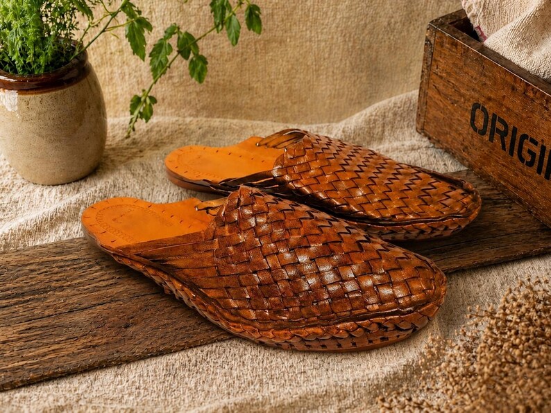 May include: A pair of brown leather woven slippers with a light brown interior. The slippers are displayed on a wooden plank, with a neutral-toned background. The slippers are a slip-on style, perfect for indoor or outdoor wear.