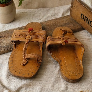 Könnte beinhalten: Ein Paar handgefertigte Ledersandalen in hellem Braun. Die Sandalen weisen aufwendige Nähte und geflochtene Details auf, mit einem kleinen roten Akzent. Das Design umfasst eine Zehenschlaufe und einen Riemen über dem Fuß.