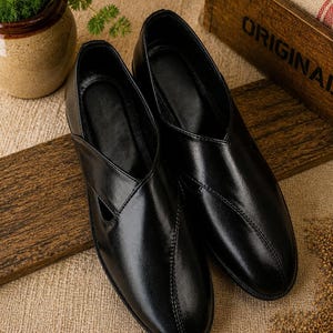May include: A pair of black leather slip-on shoes with a V-shaped design on the top. The shoes are displayed on a wooden surface with a neutral-colored fabric background. The shoes have a glossy finish and a stitched seam.