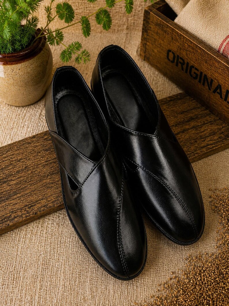 May include: A pair of black leather slip-on shoes with a V-shaped design on the top. The shoes are displayed on a wooden surface with a neutral-colored fabric background. The shoes have a glossy finish and a stitched seam.