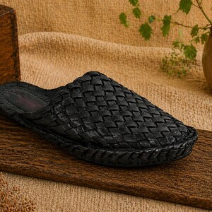 May include: A black woven leather slipper sits on a wooden surface. The slipper has a textured, basket-weave design. The background includes a wooden box, a small potted plant, and a tan fabric.