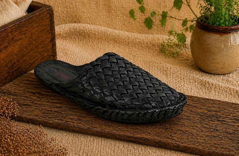 May include: A black woven leather slipper sits on a wooden surface. The slipper has a textured, basket-weave design. The background includes a wooden box, a small potted plant, and a tan fabric.
